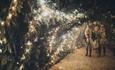 Two people walking through woodland set lit with fairy lights