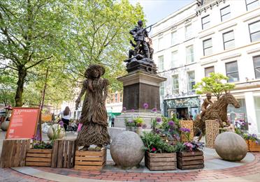 Wooden sculptures around a monument in manchester with floral displays