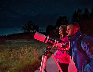 A couple standing by a telescope stargazing into the night sky in Davagh Forest at OM Dark Sky Park and Observatory.