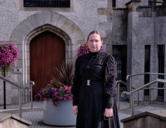 Image of woman standing under the stone arch of the Bridewell, Magherafelt.  She is dressed in a long black dress from 1800's and holds a large brass