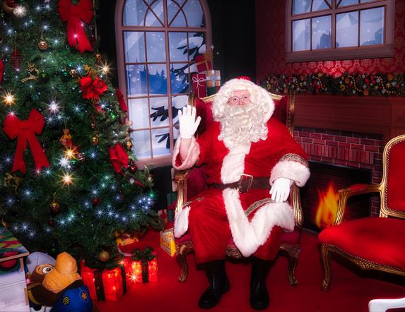 Santa sitting in his decorated grotto beside a sparkly Christmas tree with glowing presents underneath and a red and gold empty chair beside him.