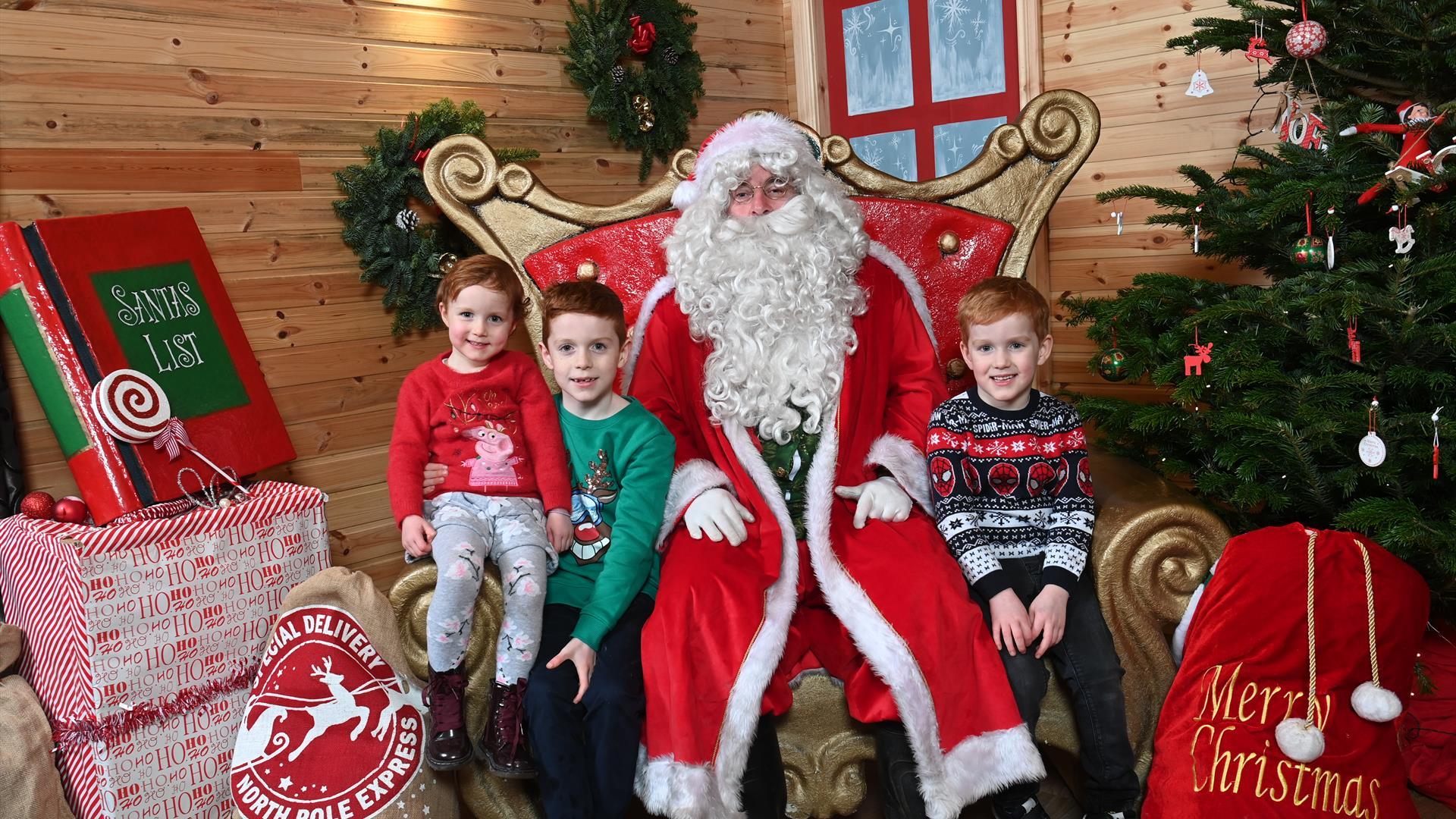 Three children visiting Santa in his grotto wearing their sparkly Christmas jumpers, surrounded by a Christmas tree and sacks of presents.