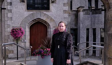 Image of woman standing under the stone arch of the Bridewell, Magherafelt.  She is dressed in a long black dress from 1800's and holds a large brass