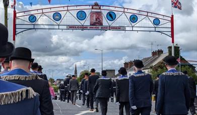 Orange men in suits and bowler hats marching under large arch