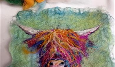 A small piece of felt craft using orange and purple hews to create an image of a highland cow's head against a moss green background.