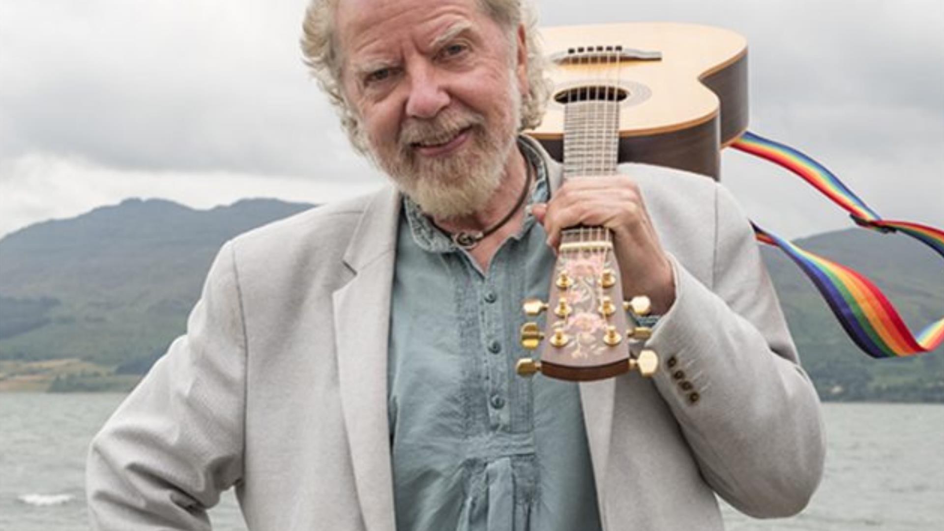 Image of Tommy Sands with a guitar over his shoulder, standing at the shore of a body of water, with mountain scenery in the background.