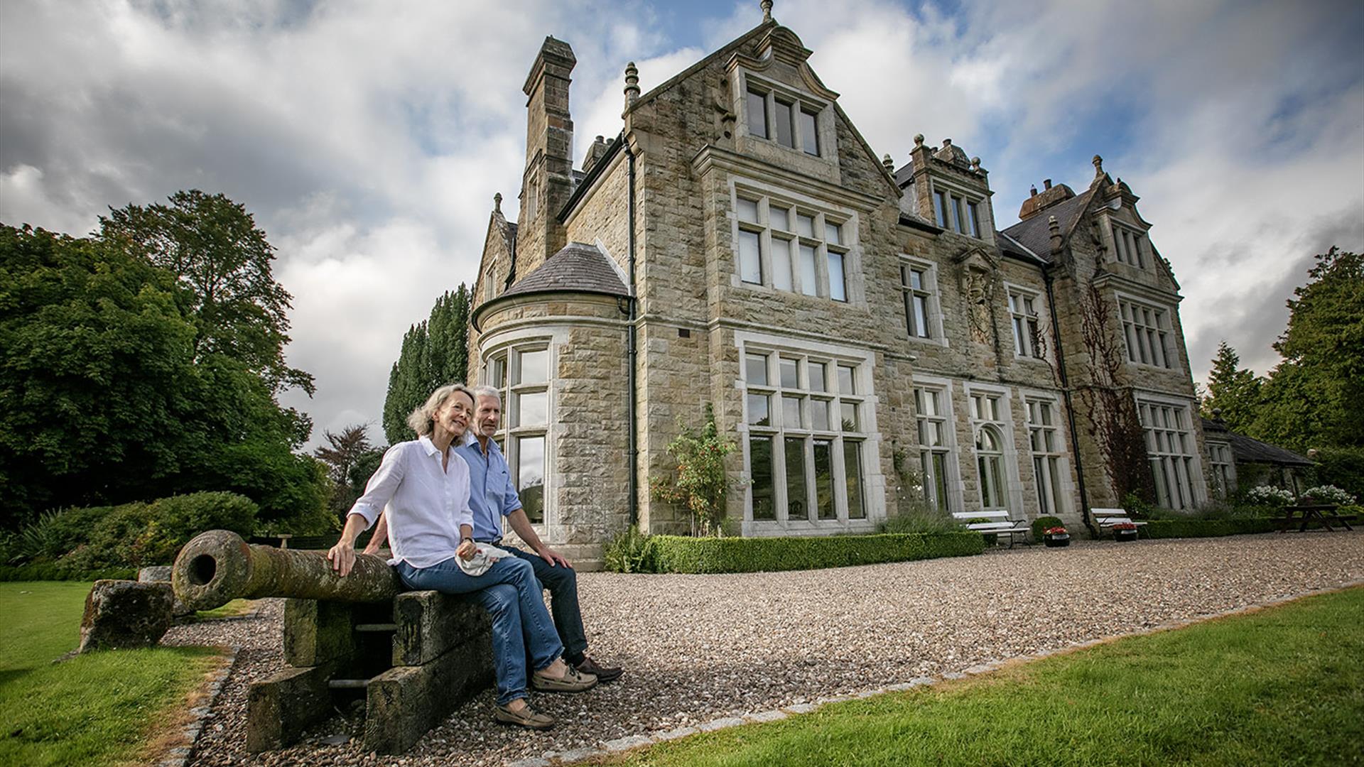 A couple sitting on an old canon in front of a large 17th century manor house