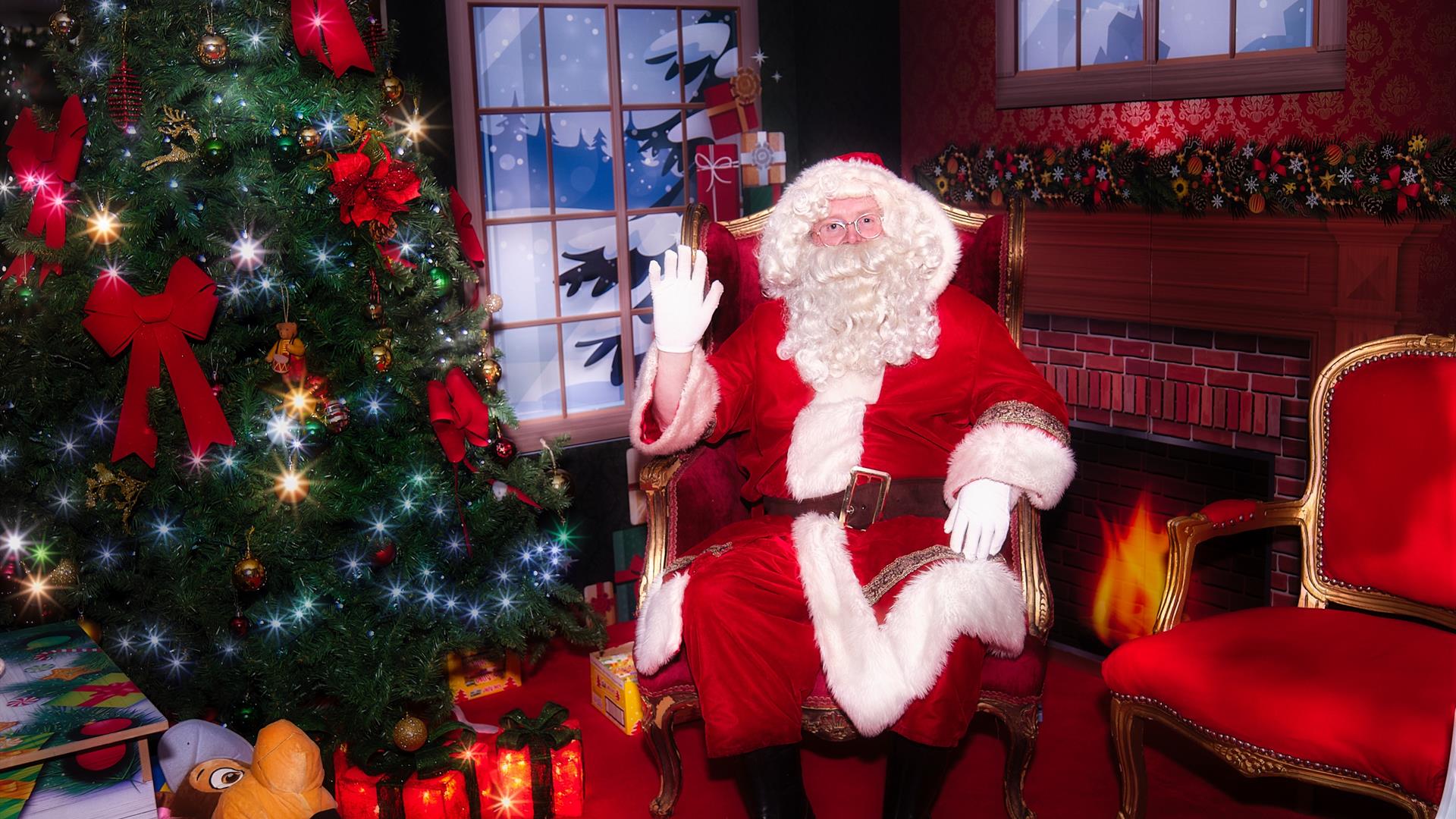 Santa sitting in his decorated grotto beside a sparkly Christmas tree with glowing presents underneath and a red and gold empty chair beside him.