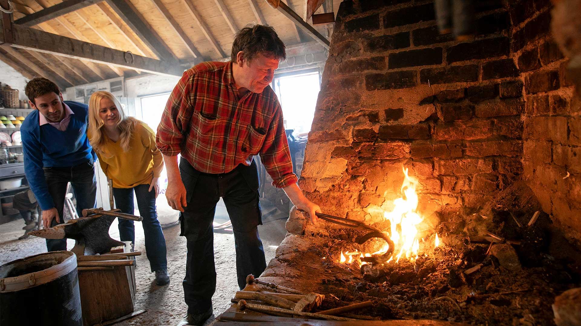 A blacksmith at the fire with a couple looking on