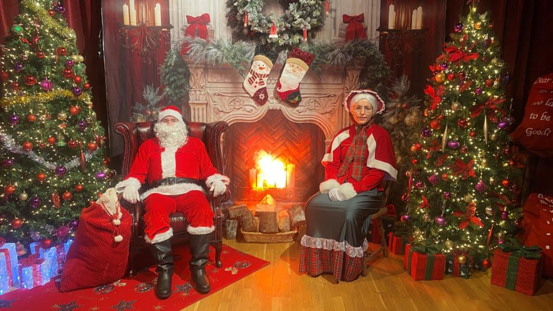 Image of Santa and Mrs Claus sitting by a fireplace surrounded by Christmas decorations
