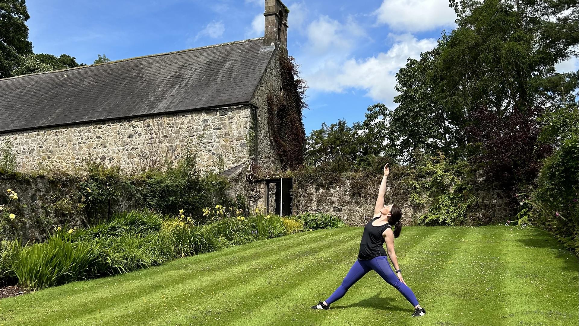 Image of a lady in purple lycra leggings and black vest top in a yoga pose with her legs standing stretched apart and one arm reaching upwards with he