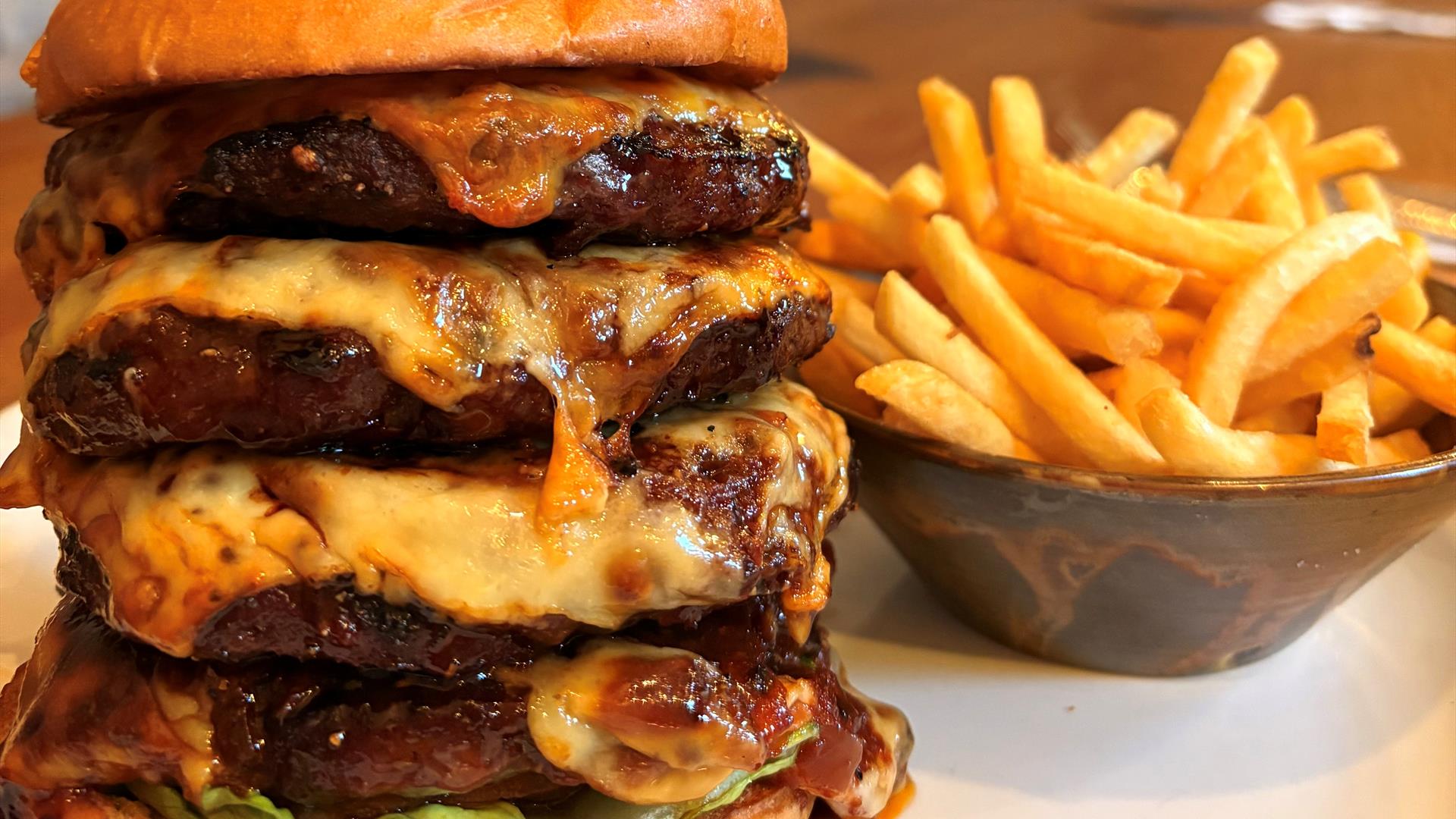 4 stacked meaty burgers in a brioche bap with smoked cheese and bacon and a bowl of fries