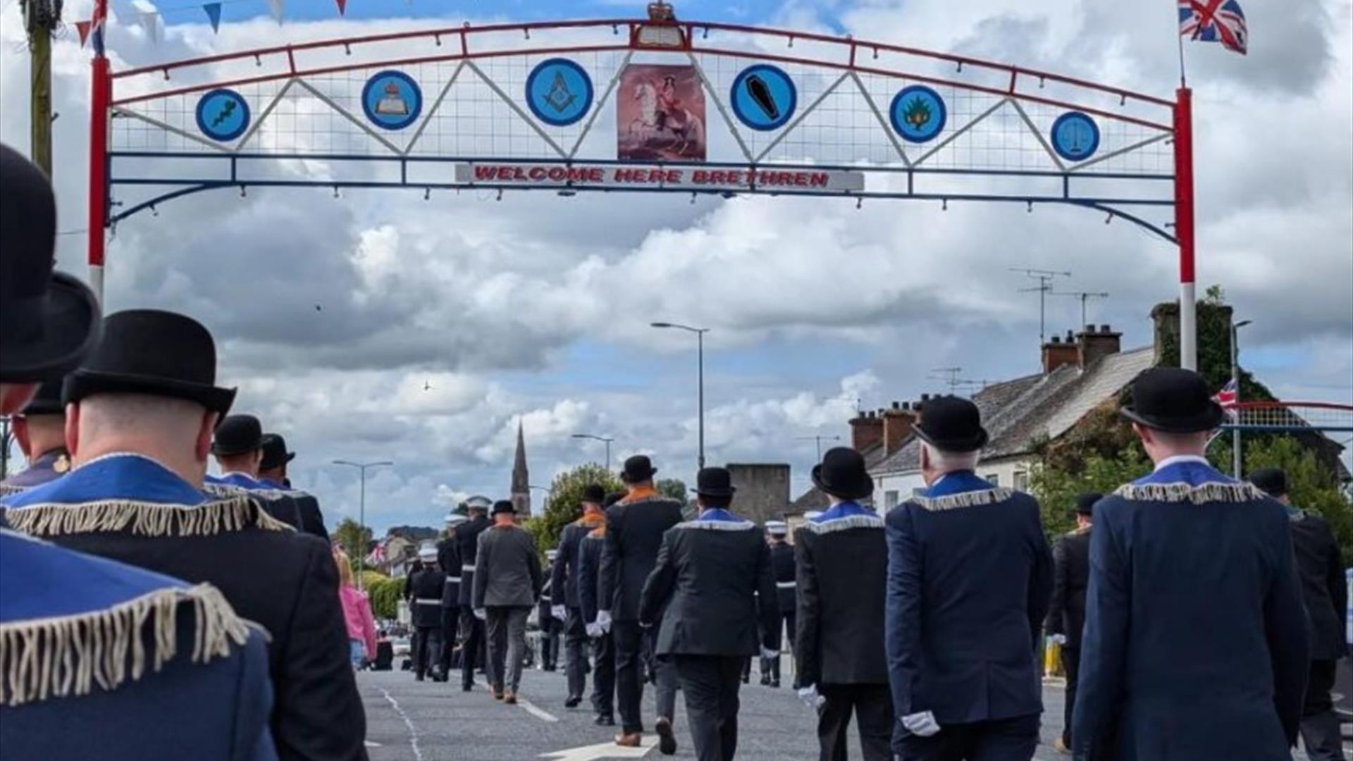 Orange men in suits and bowler hats marching under large arch