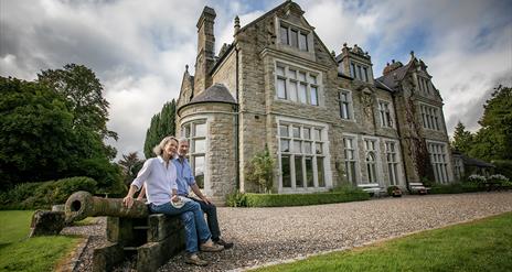 A couple sitting on an old canon in front of a large 17th century manor house