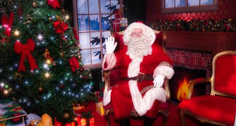 Santa sitting in his decorated grotto beside a sparkly Christmas tree with glowing presents underneath and a red and gold empty chair beside him.