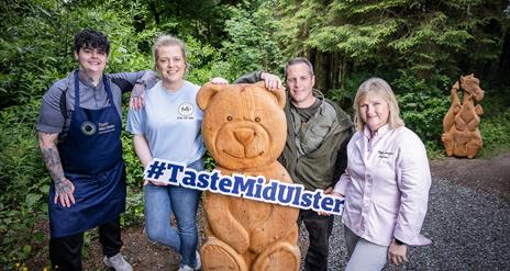 Chef Lauren Shimmon wearing navy Taste Mid Ulster apron, with her elbow resting on Bethany Boyd, Betty's Ice Cream leaning on a wooden teddy bear stru