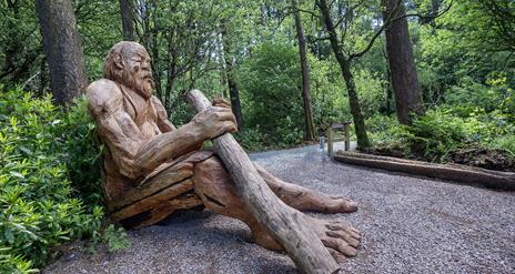 Large wooden sculpture of a giant sitting on the ground leaning against a tree holding a big stick.