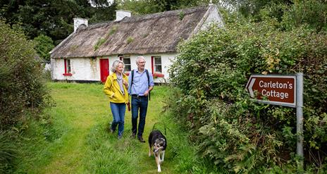 A mature couple and their dog walk through grass away from an Irish thatched cottage.