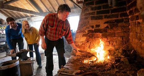 A blacksmith at the fire with a couple looking on