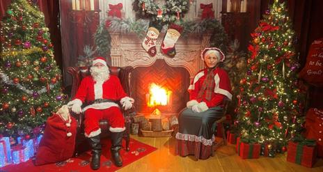 Image of Santa and Mrs Claus sitting by a fireplace surrounded by Christmas decorations