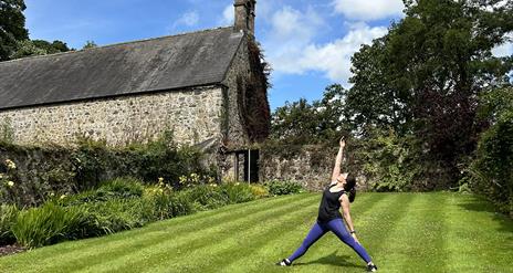 Image of a lady in purple lycra leggings and black vest top in a yoga pose with her legs standing stretched apart and one arm reaching upwards with he