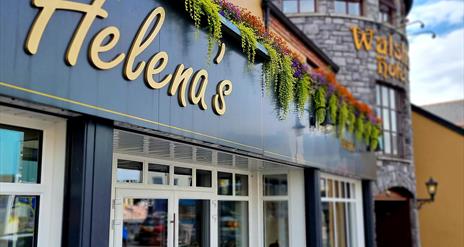 Exterior of Helena's Coffee Shop and Bistro, featuring a modern black and white facade with gold lettering. Colorful hanging plants decorate the top o