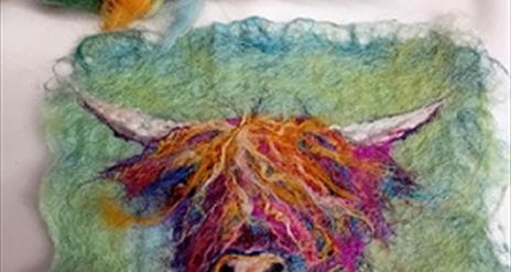 A small piece of felt craft using orange and purple hews to create an image of a highland cow's head against a moss green background.