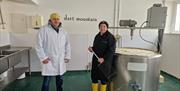 Two people in protective clothing standing beside a large stainless‑steel cheese‑making vat inside a dairy facility, with “Dart Mountain” on the wall