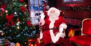 Santa sitting in his decorated grotto beside a sparkly Christmas tree with glowing presents underneath and a red and gold empty chair beside him.