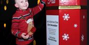 Young boy wearing a red reindeer jumper posting his letter to Santa in a large red Santa's Post box with snow flakes over it