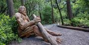 Large wooden sculpture of a giant sitting on the ground leaning against a tree holding a big stick.