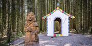 A wooden structed of the witch from Hansel and Gretel standing in front of a small white hut with images of sweets painted on it.  in the background i