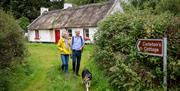 A mature couple and their dog walk through grass away from an Irish thatched cottage.