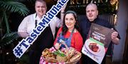 3 people standing together  holding a large “#TasteMidUlster” sign, a wicker basket filled with food and fresh greens, and a promotional poster for Mi