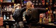 Two bartenders in black uniforms are seen from behind as they prepare drinks at Jack's Bar. Shelves filled with various liquor bottles, including gin