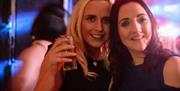 Two women smiling and posing for a photo at Jack's Bar. One is holding a drink in her hand, and the background features colorful lights, creating a li