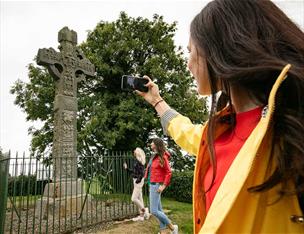 Two girls stand by Ardboe Cross as another woman photographs them on her phone. 