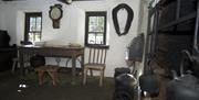 Image of the inside of a cottage with wooden table and chairs, old kettles and an old clock