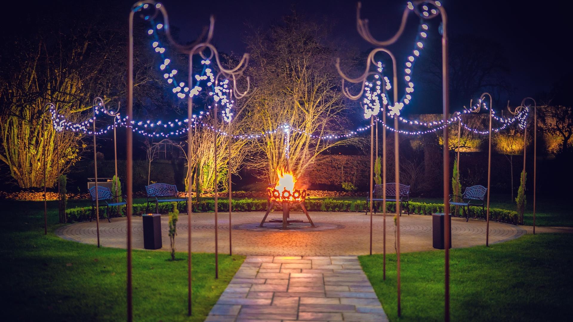 Paved path leading to circular patio with fairy lights and fire pit. Paved path leading to circular patio with fairy lights and fire pit.