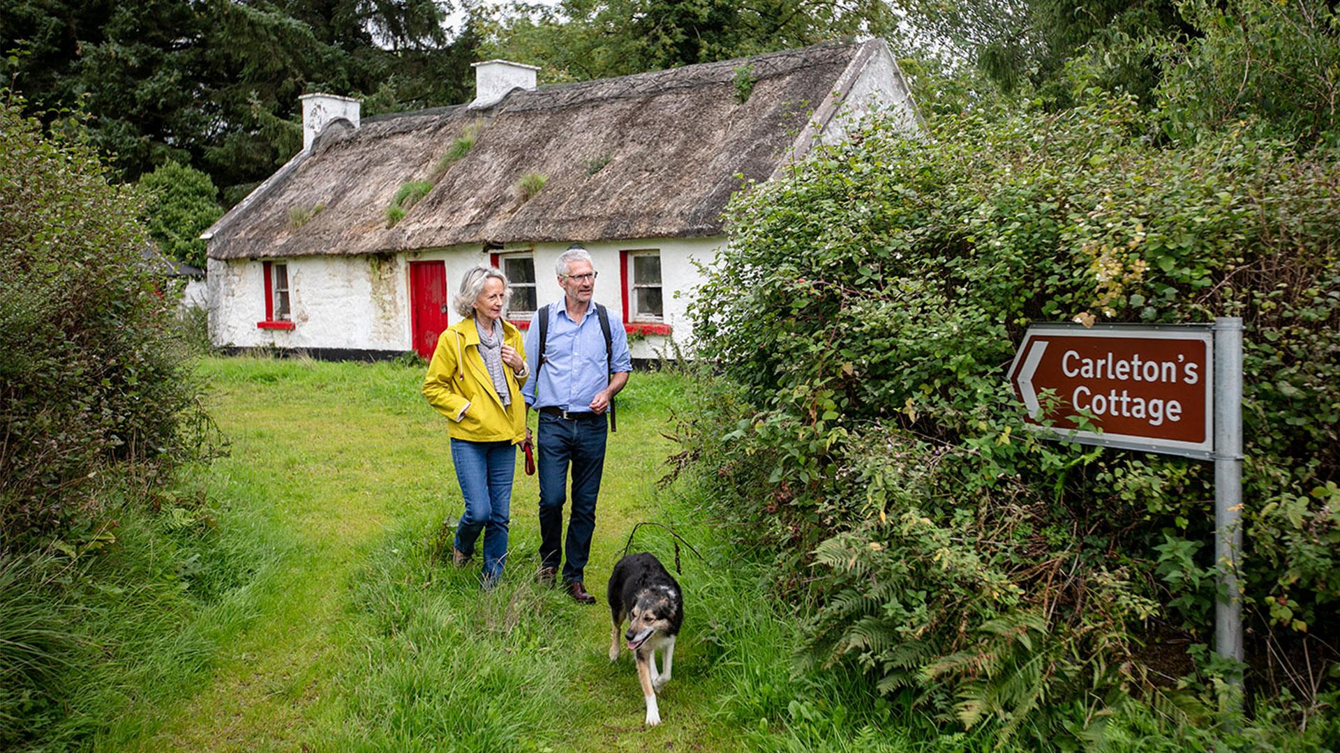 A mature couple and their dog walk through grass away from an Irish thatched cottage.
