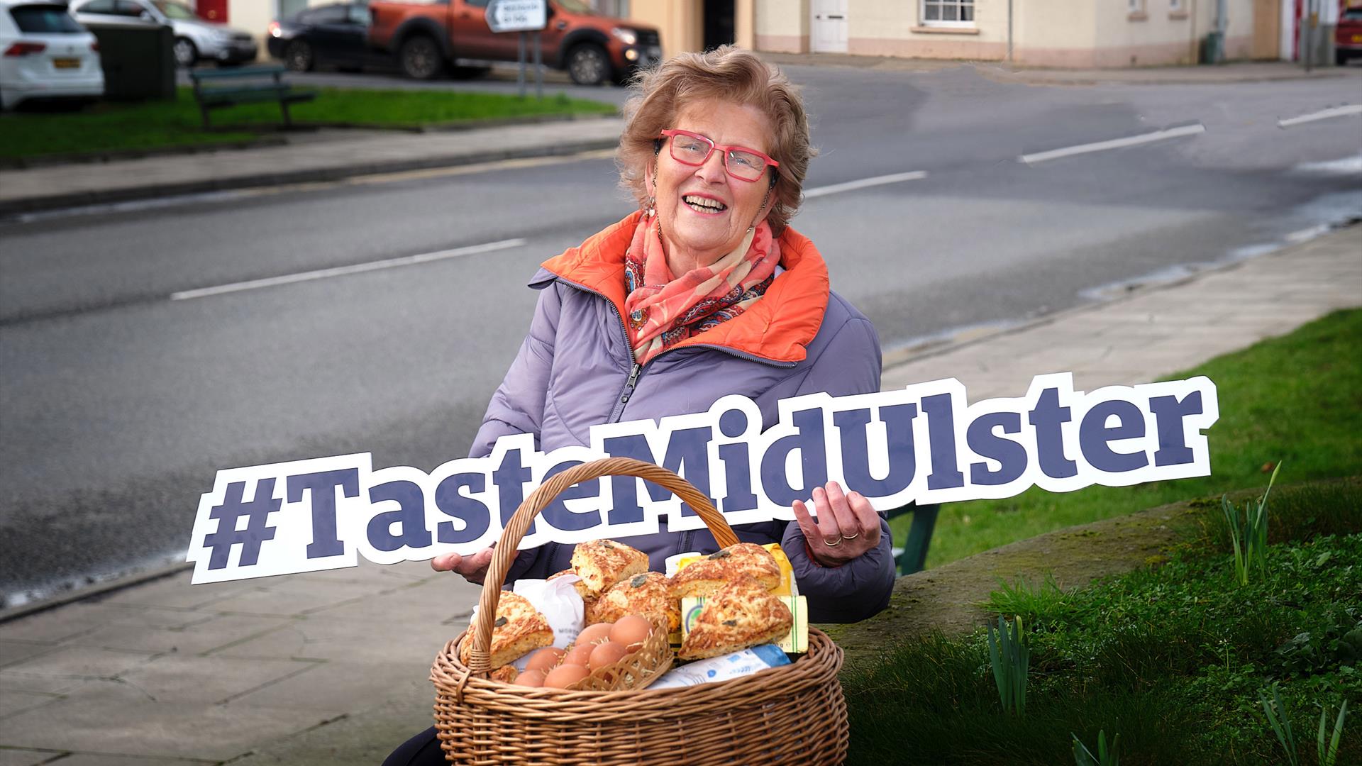 Norah Brown MBE in Moy Village holding a Taste Mid Ulster sign.
