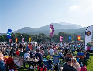 Eats and Beats festival with vibrant flags, food stalls, and a lively crowd enjoying music, food, and drinks under sunny skies, set against a scenic backdrop of Slieve Donard mountain, the highest mountain in Northern Ireland.