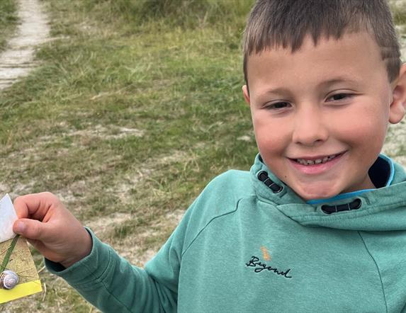 Child standing at Tyrella holding artwork he made with paper, shells, grass and sand.