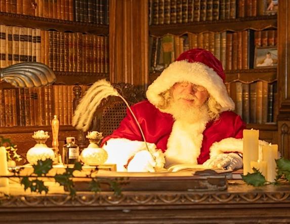 Santa in his red suit sitting at his desk in his library, with walls filled with books writing with a quill.