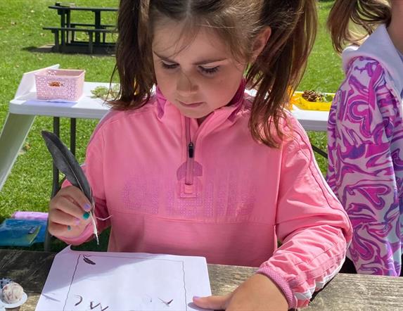 A girl standing at a table surrounded by the green grass of the Park, writing with a quill.