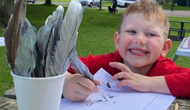 A boy sitting at a table smiling whilst writing with a quill