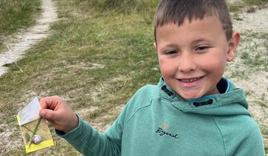 Child standing at Tyrella holding artwork he made with paper, shells, grass and sand.