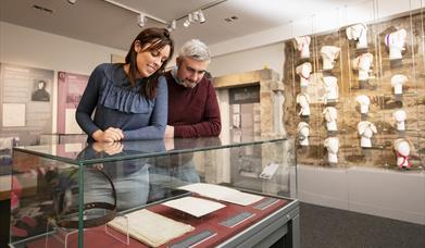 A man and a woman look at archives related to the old gaol in Downpatrick, with a museum display of bonnets in the background