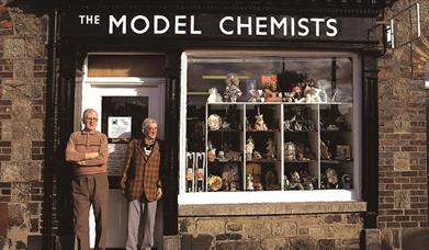 Two gentlemen standing outside 'The Model Chemist' building as part of the Bessbrook Historic Model Village