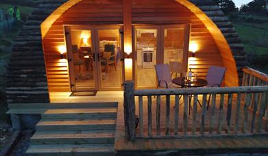 exterior veiw of Log cabin ,  4 wide wooden steps up to patio veranda. Round patio table and 2 chairs on veranda with cabin light up behind.
